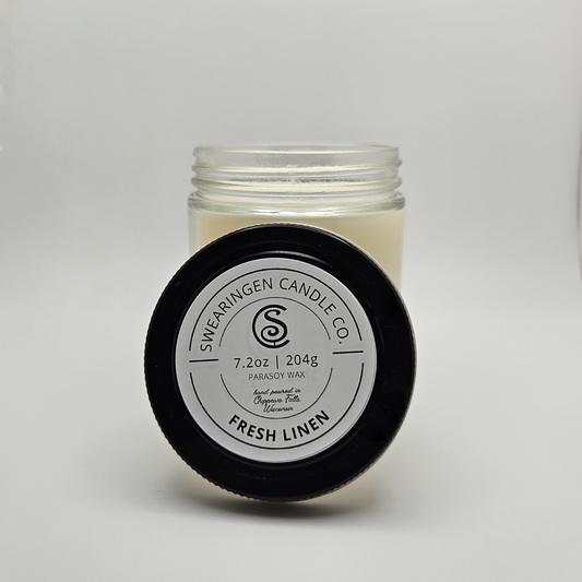 Fresh Linen hand-poured parasoy candle in a clear glass jar with a black lid, shown in a three quarter view - Swearingen Candle Co.